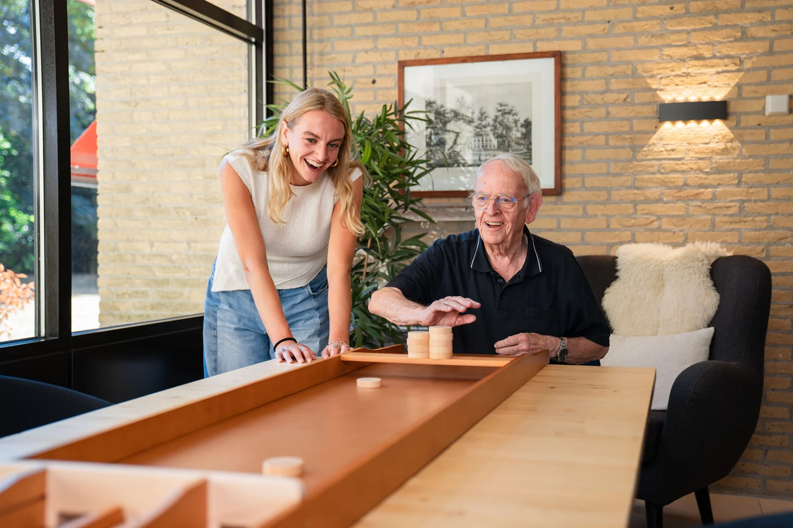 Een jonge gezelschap aanbieder speelt sjoelbak met een senior man in de woonkamer